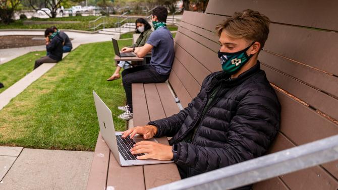 Young man wearing a Cal Poly face mask while typing on a laptop