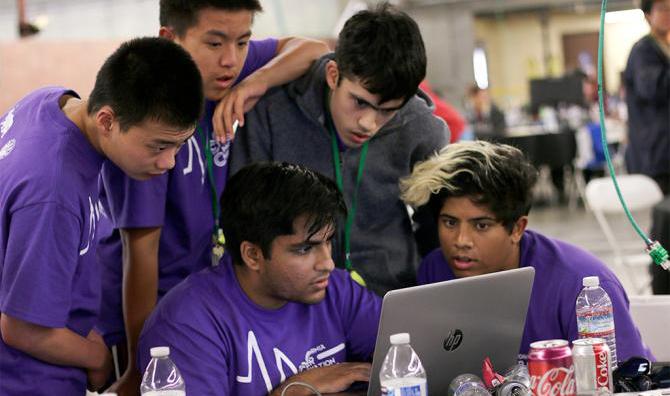 A group of high school students lean over a laptop screen as one of them types