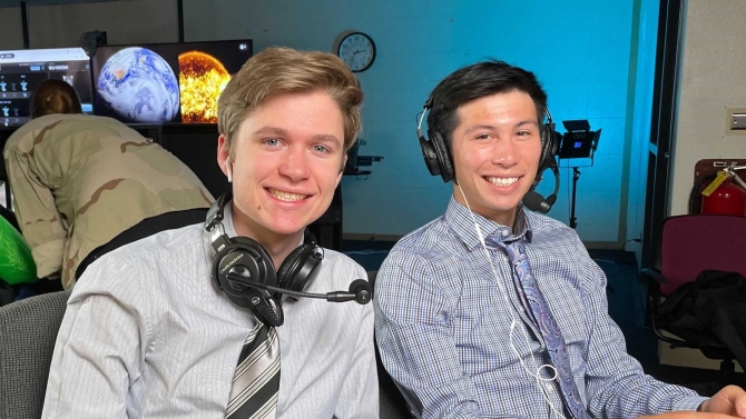 two broadcast anchors smiling in front of a wooden desk
